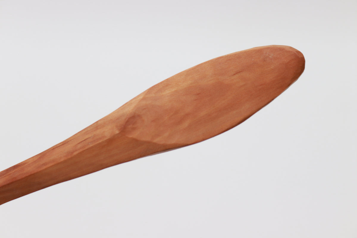 The handle of a wooden spoon, shown on a white background. The forward portion of the handle is tapered so that it's in the form of two sloped faces that meet in a distinct raised midline. However, the rear portion of the handle is a separate surface of its own, tapered toward the front but otherwise oval. "Take me in your hand," it seems to whisper, "Take me in your hand and do what you want with me."