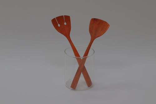 a pair of cherry salad servers, 10 inches tall and 3 inches wide at their business end, are leaning casually in a glass cylinder in front of a white/gray background.