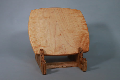 Light caramel colored with reddish highlights curly maple cutting board on a stand against a gray background. Grain runs top to bottom of picture.