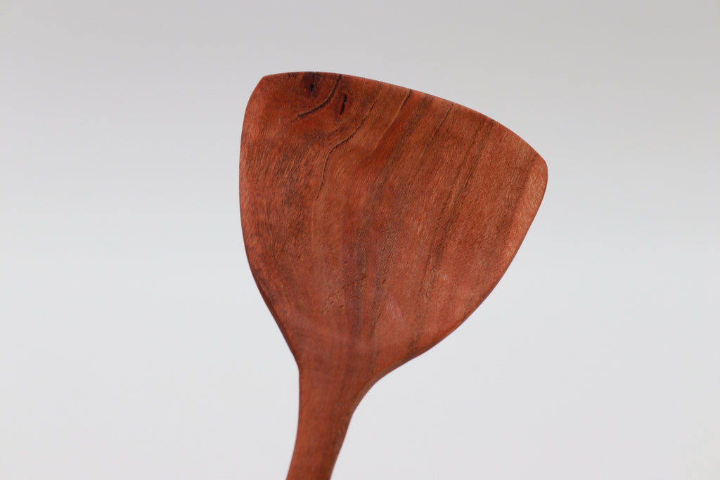 A gorgeous piece of cherry wood, shaped into a spatula-like salad server, leans jauntily towards the right in front of a white background.